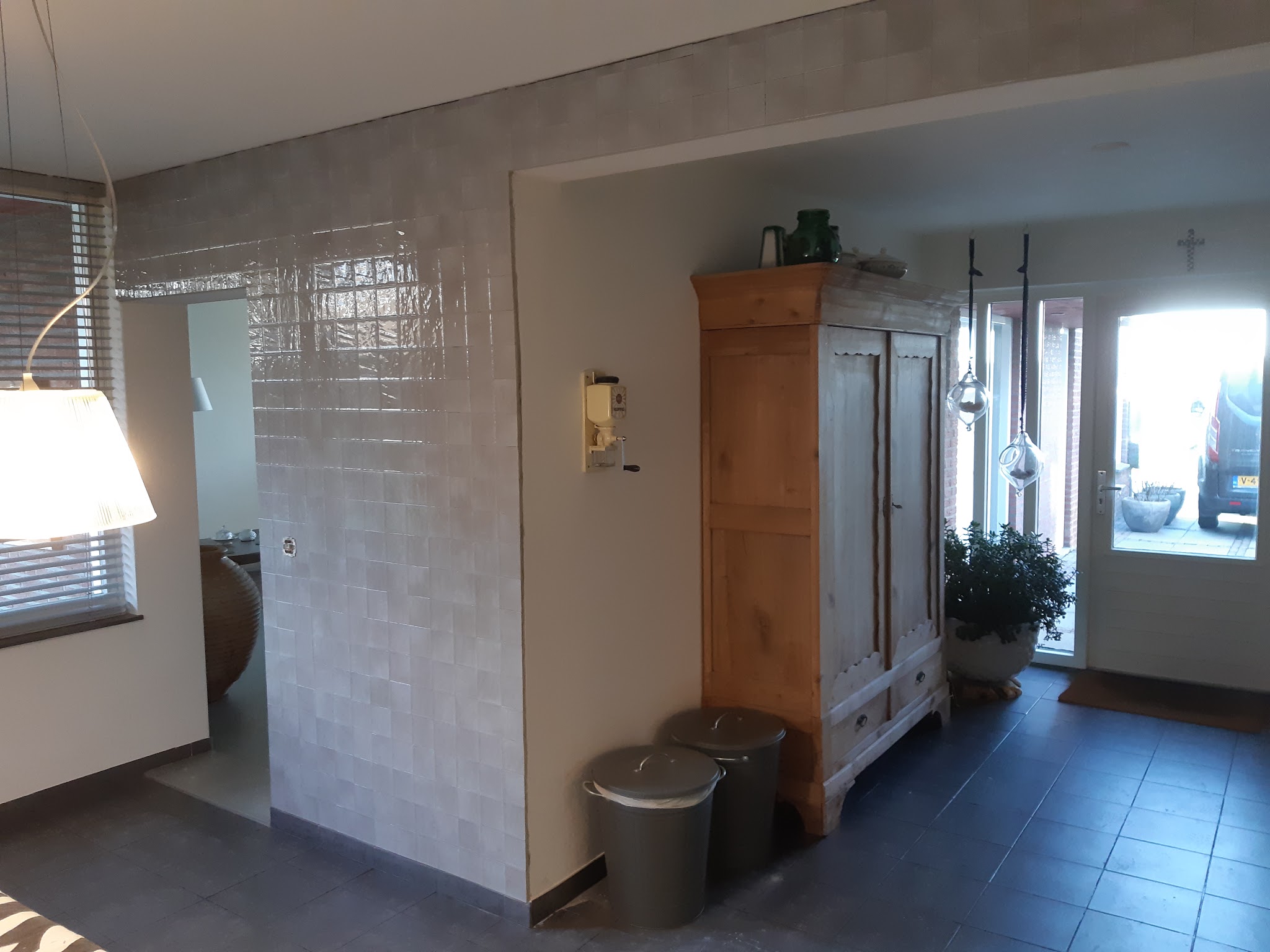 Een hal met een glanzend betegelde wand in handvormtegels en een donkere leisteenvloer